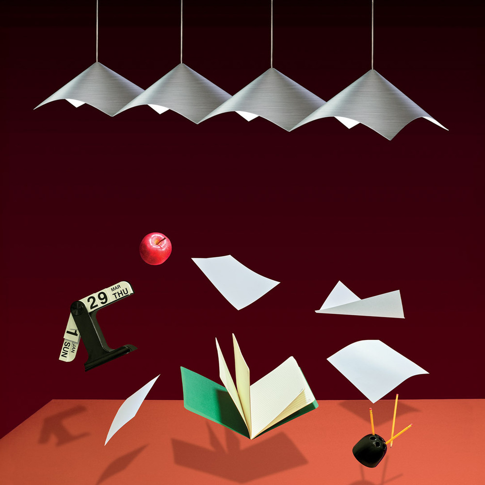 Illuminated Iperbole suspension lamps on a burgundy background, with a book, sheets of paper, a calendar, an apple and pencils suspended above an orange surface.