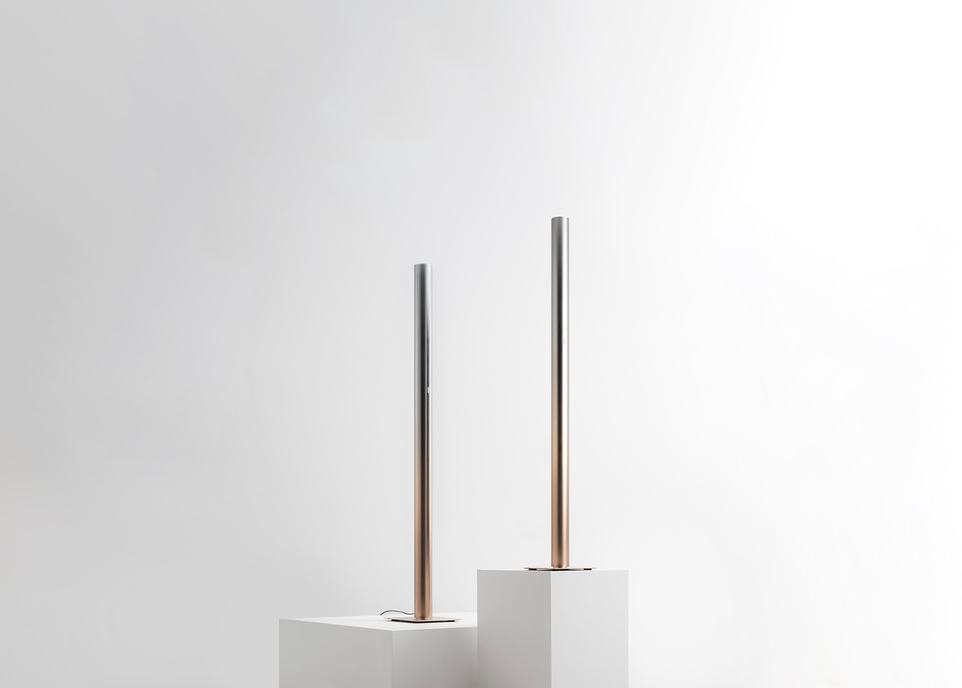 Two slim floor lamps with slender cylindrical stems, silver top graduating to rose gold base, displayed on white pedestals