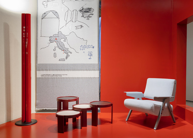 Allestimento della mostra per i 100 anni di Gianfranco Frattini con lampada Megaron rossa, tavolini e poltrona di design su sfondo rosso.