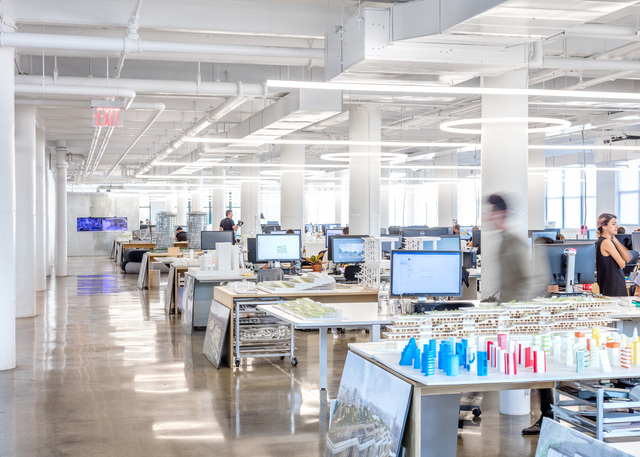 Uffici di BIG a New York in un ampio open space con tavoli di lavoro, computer, modelli architettonici e illuminazione lineare a soffitto.