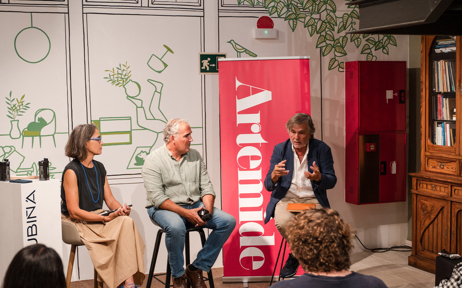 Tre persone sedute in conversazione a un evento del marchio Artemide, con un banner rosso Artemide visibile sullo sfondo.
