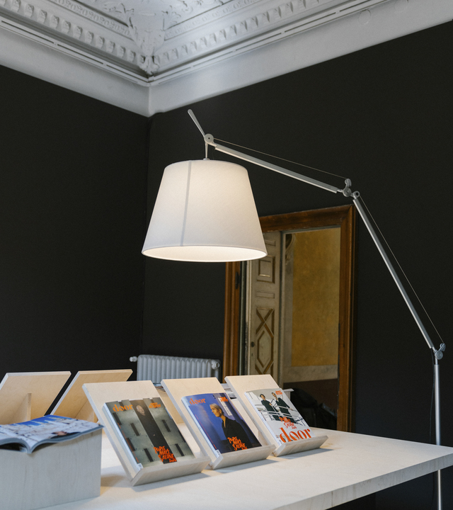 Lampada da terra Tolomeo con diffusore acceso illumina un tavolo espositivo con riviste door, in un interno con pareti scure e soffitto decorato.