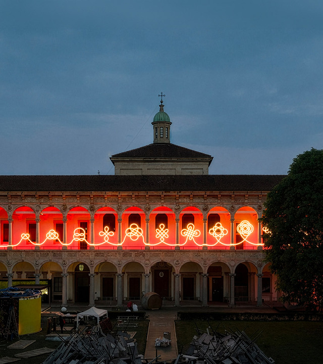 Facciata del chiostro dell’Università Statale di Milano al crepuscolo, con installazione luminosa lineare che disegna motivi intrecciati tra le arcate.