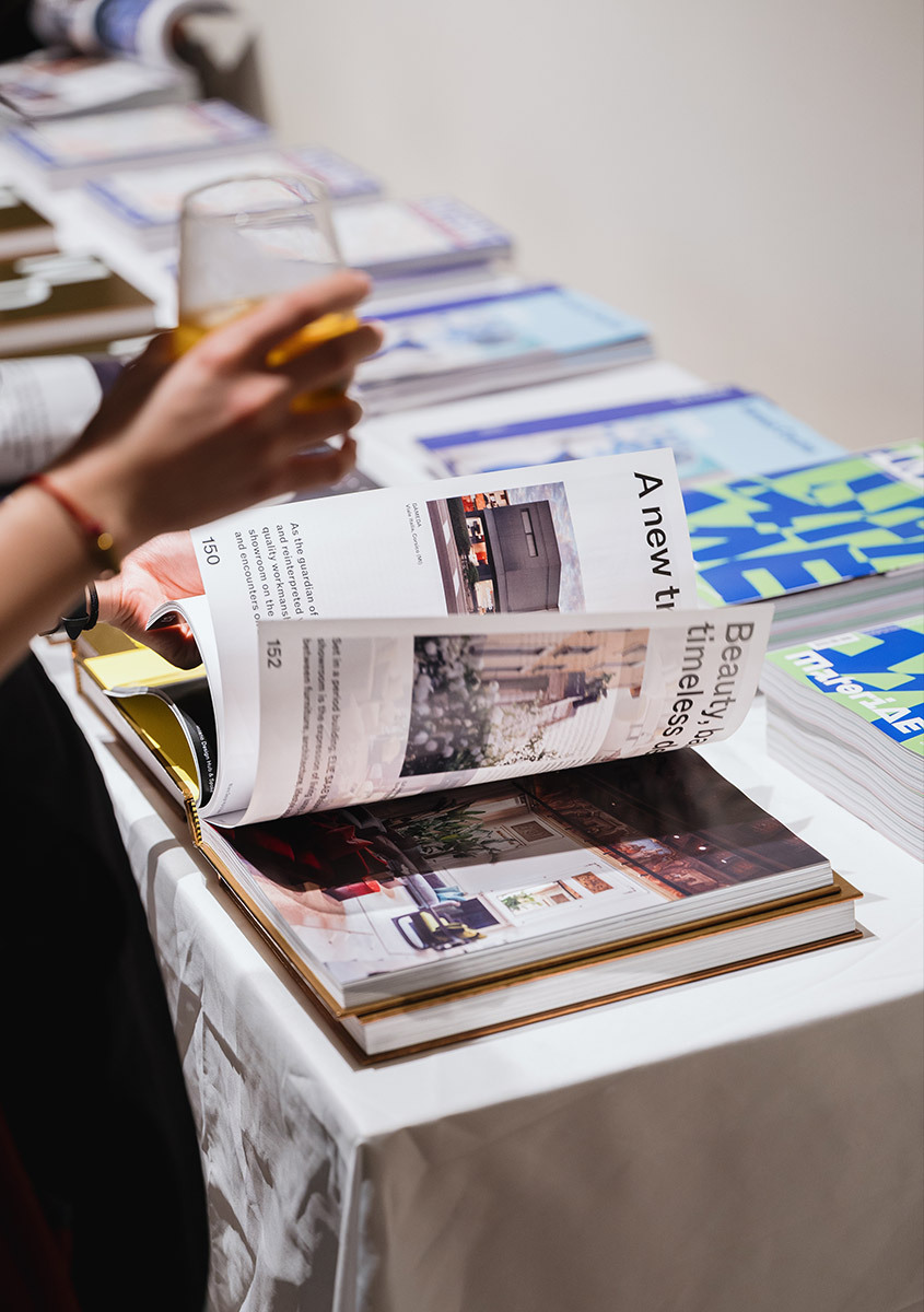 Person browsing a catalogue at an event, holding a glass of wine while flipping through pages featuring lighting products.