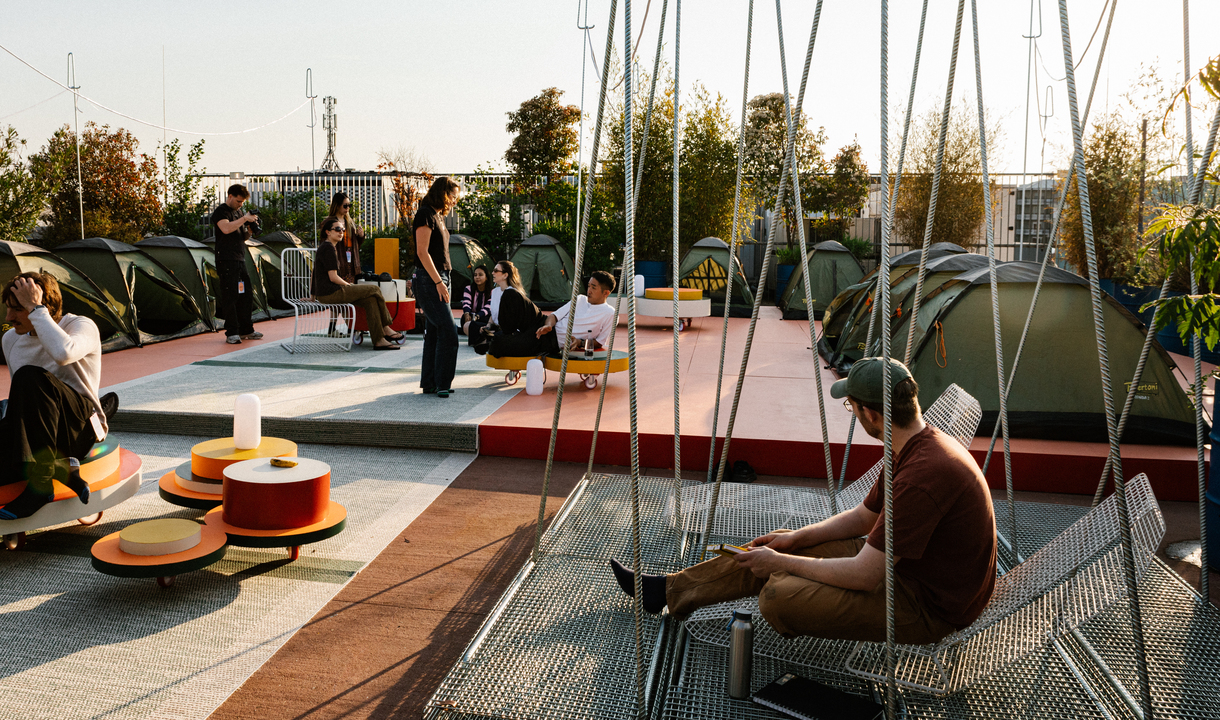 Installazione outdoor con tende, sedute sospese e arredi colorati su una terrazza, animata da persone al tramonto.