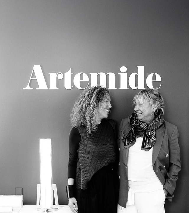 Ritratto in bianco e nero di Carlotta de Bevilacqua e Giulia Foscari in piedi sotto il logo Artemide, accanto alla lampada Criosfera.