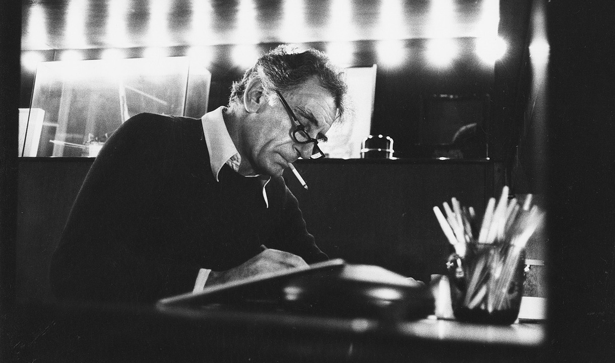 Foto storica di Gianfranco Frattini al lavoro alla scrivania, in un ritratto in bianco e nero illuminato da una fila di luci sullo sfondo.