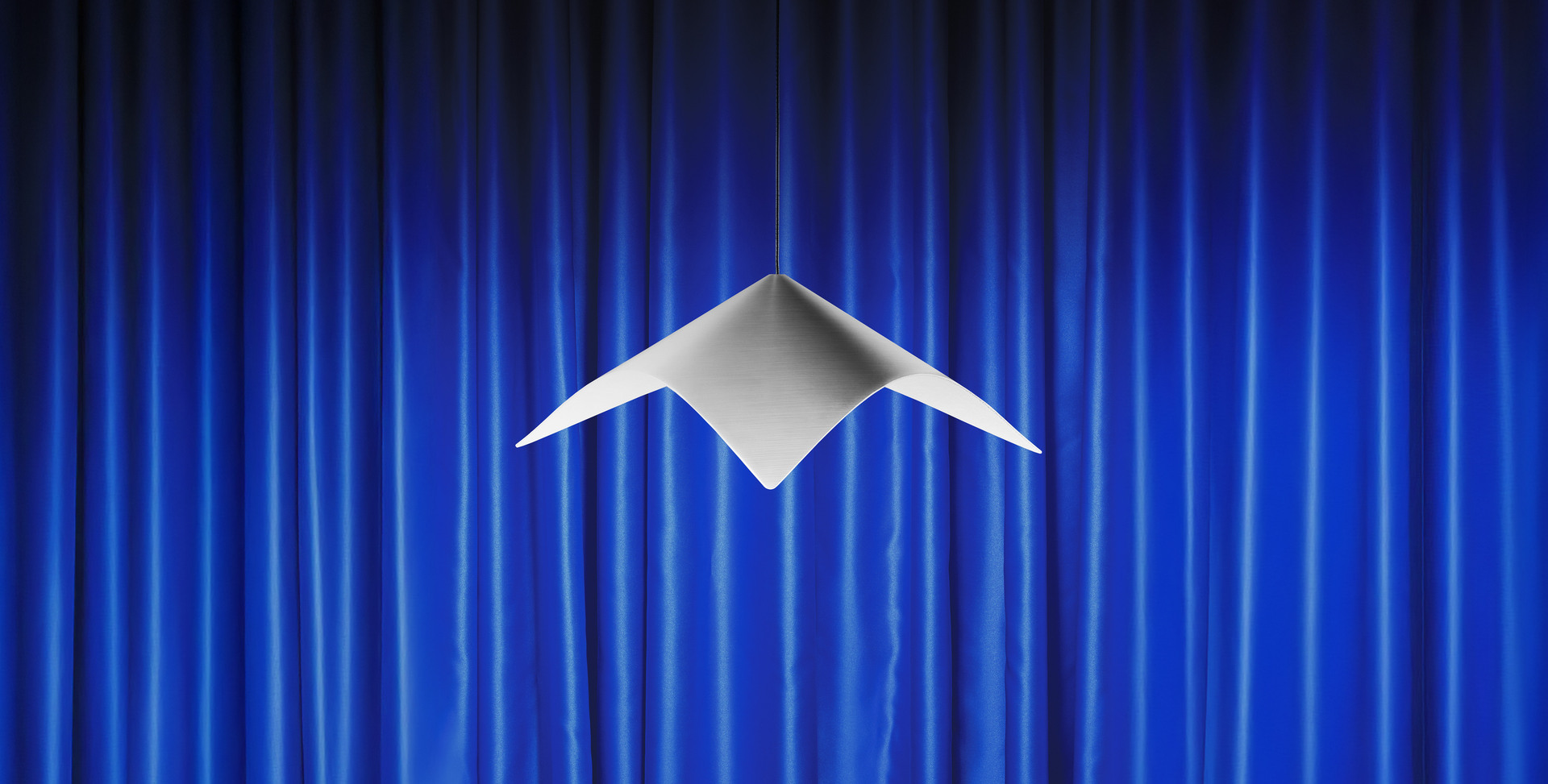 Lampada a sospensione Iperbole davanti a tende da teatro blu.