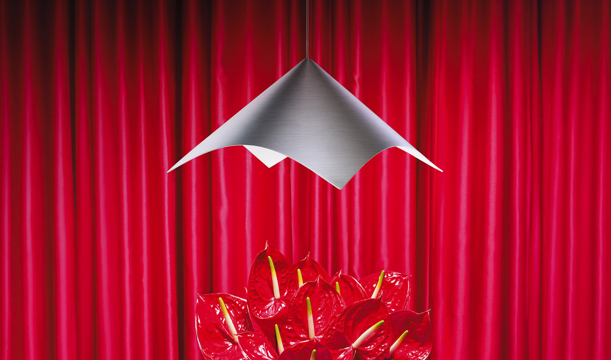 Lampada a sospensione Iperbole in metallo dalla forma scultorea, sospesa sopra un gruppo di anthurium rossi davanti a un fondale di tende rosse. Foto di Pierpaolo Ferrari.