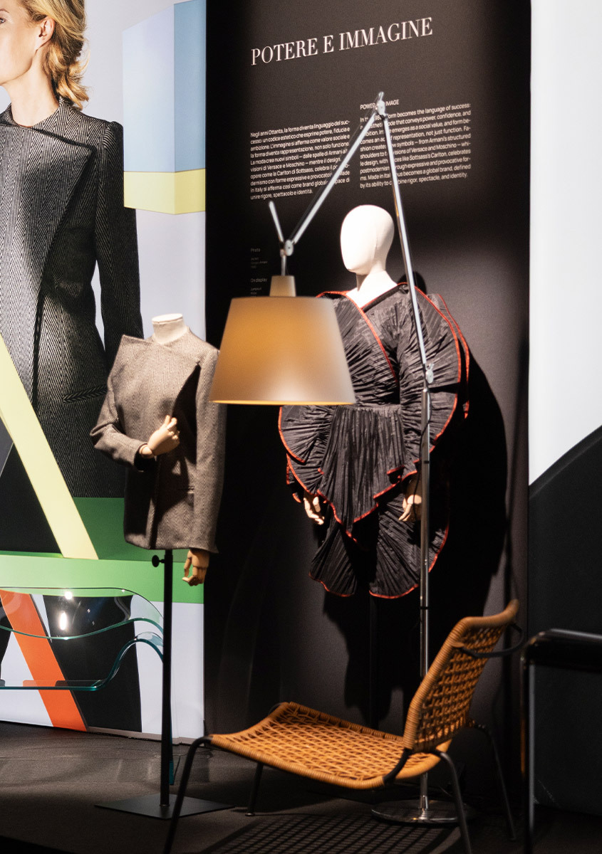 Lampada da terra Artemide Tolomeo Mega accesa, esposta accanto a un abito su manichino e a una seduta in vimini nella mostra ABITO.