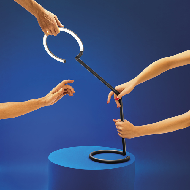 Image of the black Vine Light table lamp placed on a blue cylinder, in the scene there are arms and hands grasping it.