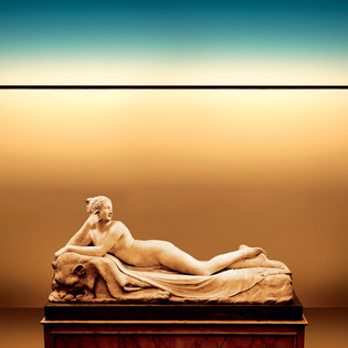 Statua classica reclinata su un piedistallo, illuminata da Dusk, una sottile linea luminosa orizzontale, in uno spazio dai toni caldi con luce gradiente. Foto di Pierpaolo Ferrari.
