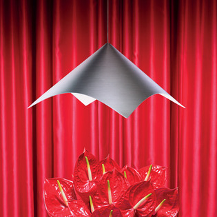 Lampada a sospensione Iperbole in metallo sopra un gruppo di anthurium rossi, davanti a un fondale di tende rosse. Foto di Pierpaolo Ferrari.