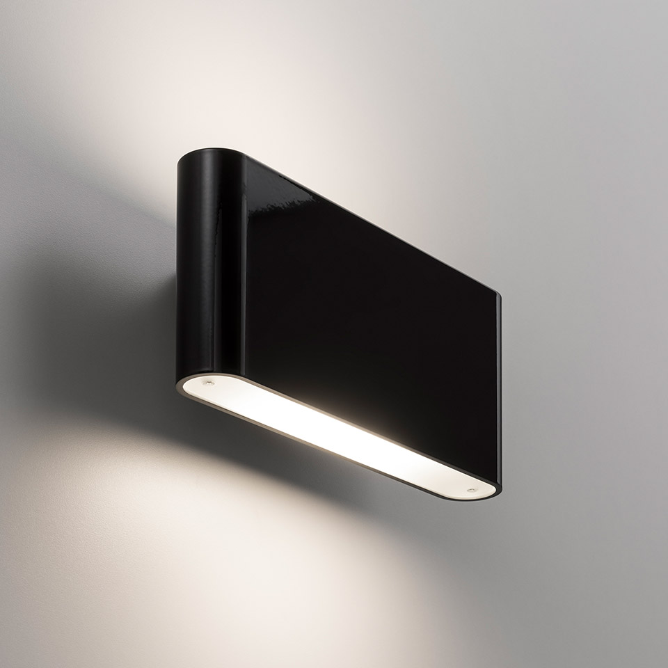 Lampada da parete Artemide Two Flags Parete nera, corpo rettangolare con diffusore inferiore a luce bianca calda, alone morbido sul muro