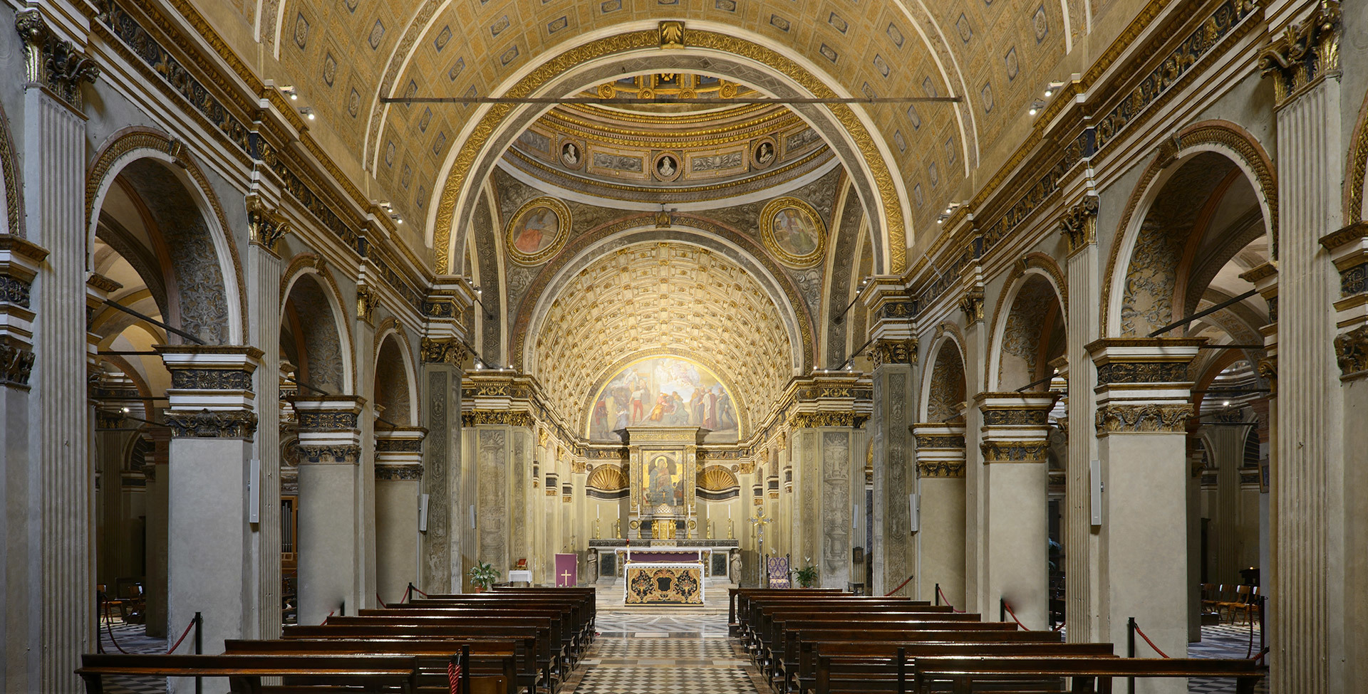 Interior of a Renaissance church with barrel-vaulted nave, gilded mosaics, marble columns, wooden pews, and illuminated apse altar.