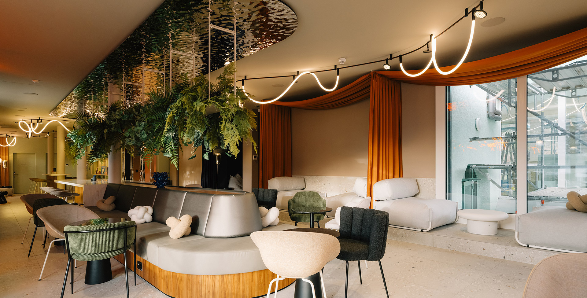 Spacious lounge with upholstered seating, hanging plants, orange curtains, and the Sylt ceiling system with luminous lines.