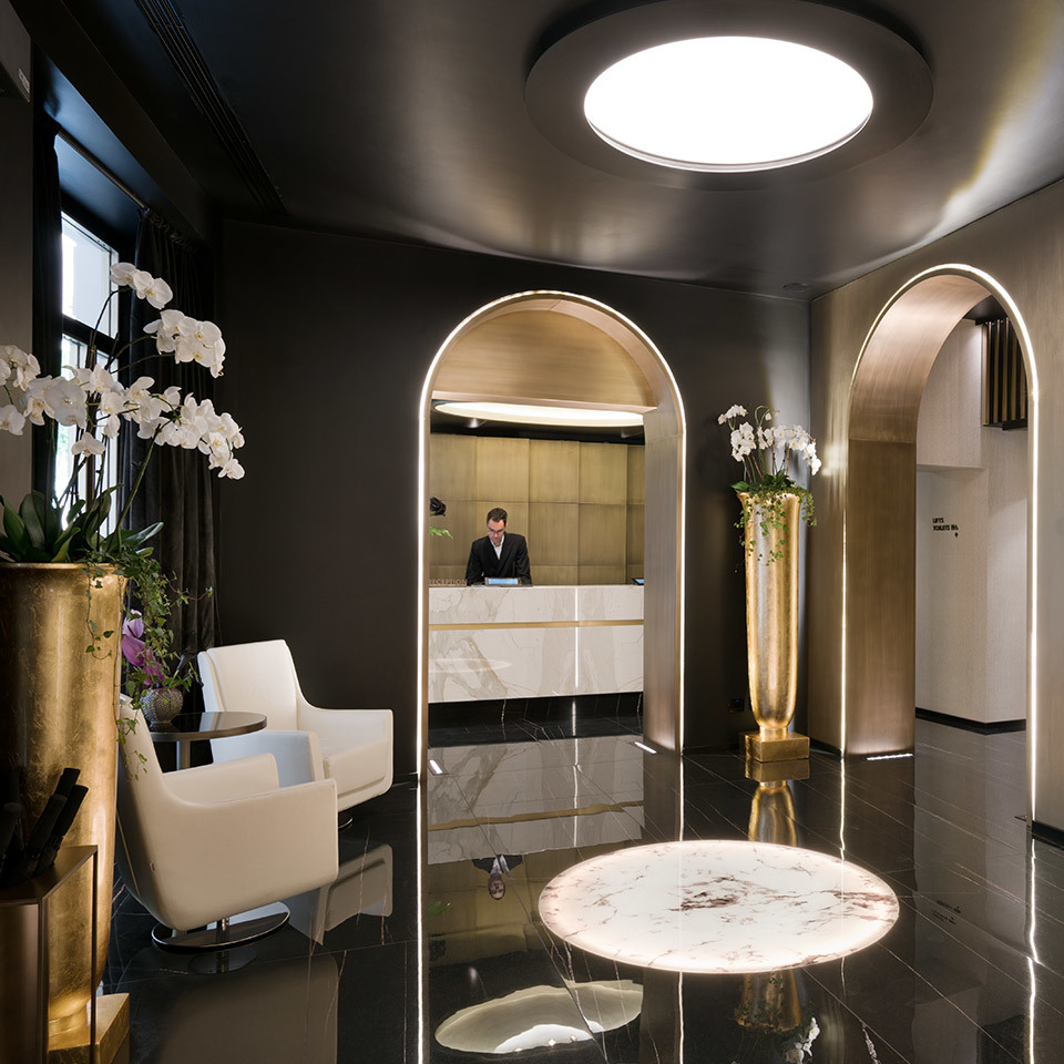 Elegant hotel lobby with dark walls, arched gold-trimmed doorways, circular ceiling light, marble floor accents, and a reception desk in background.