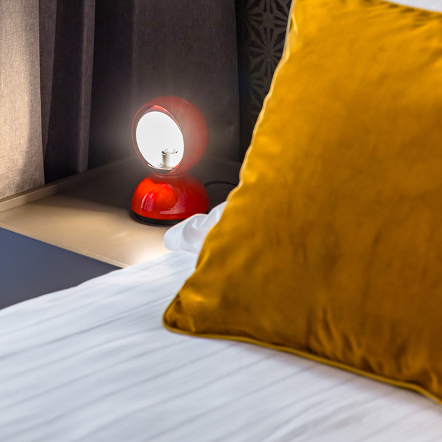 Artemide Eclisse red table lamp glowing on a bedside shelf next to white bedding and a mustard velvet pillow.