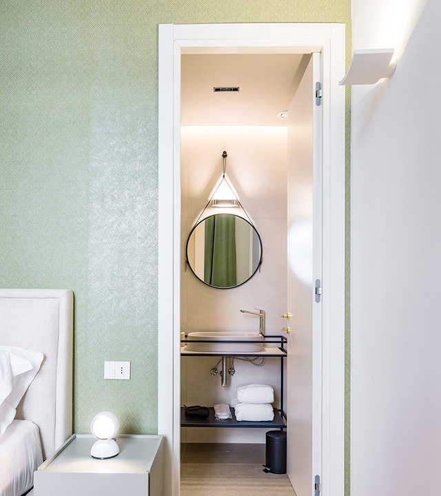 Hotel bedroom with Eclisse table lamp on nightstand, doorway revealing bathroom with pendant light above sink and round mirror.