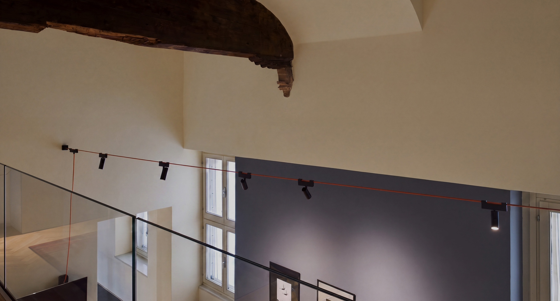 Funivia system with red cable and adjustable spotlights in a double-height space with historic wooden beams.